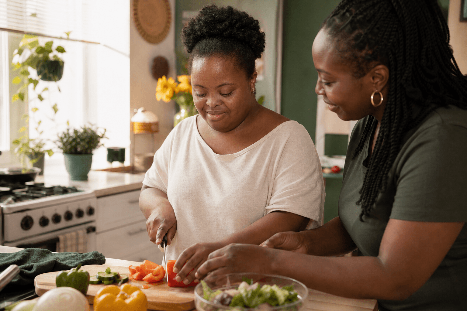 A Black woman with Down syndrome prepares vegetables in her kitchen while a support professional coaches beside her.