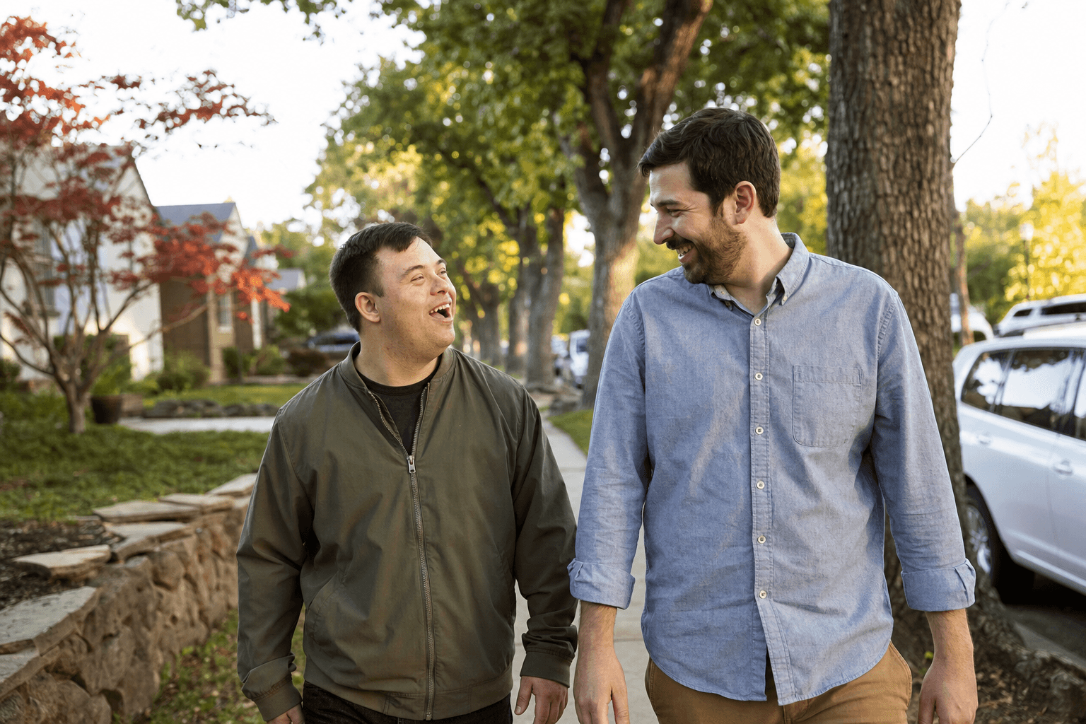 Two men walking and laughing together on a tree-lined neighborhood sidewalk in the late afternoon.